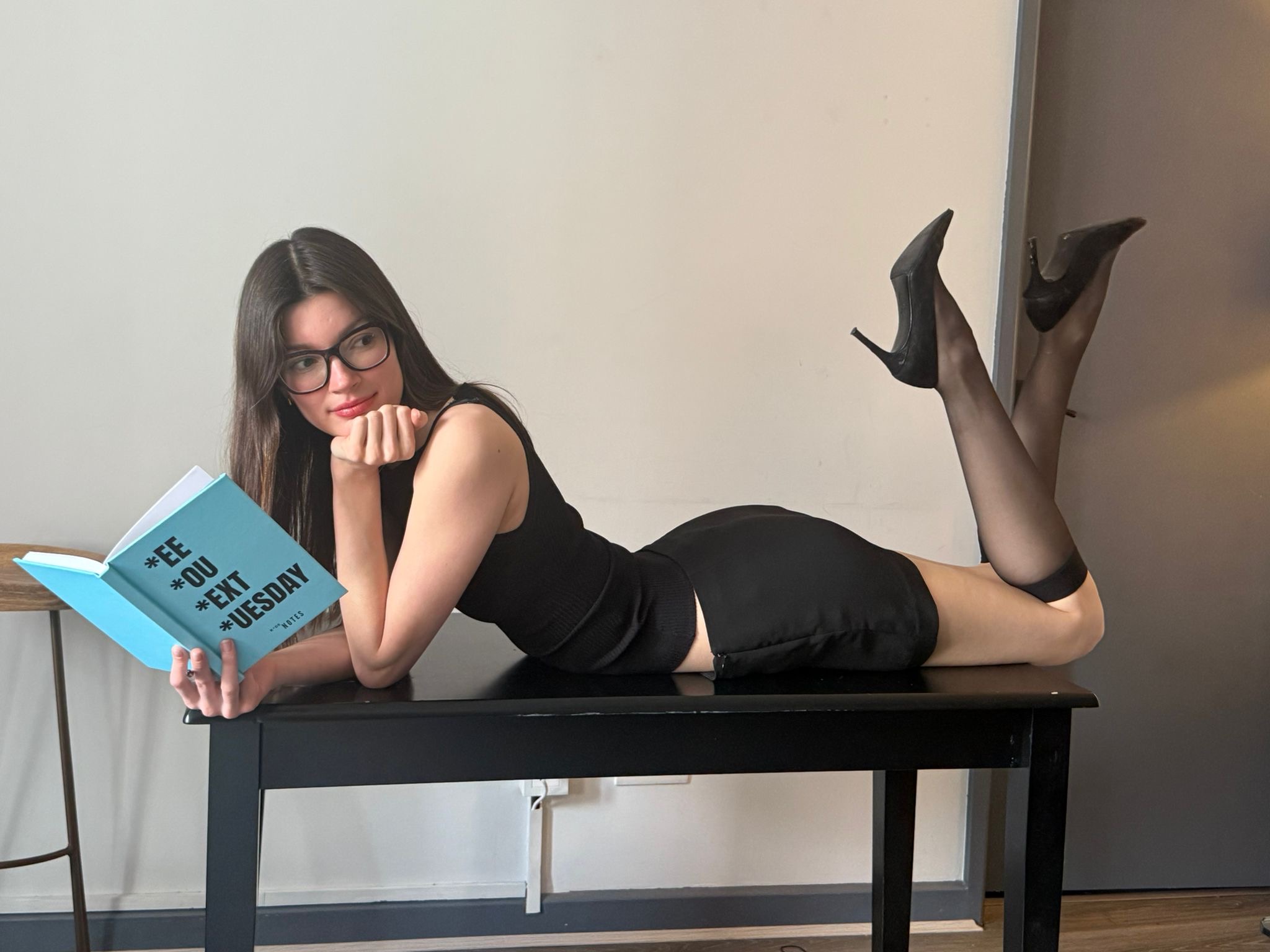 Cyan girl reclining on a black bench reading the *EE *OU *EXT *UESDAY notebook