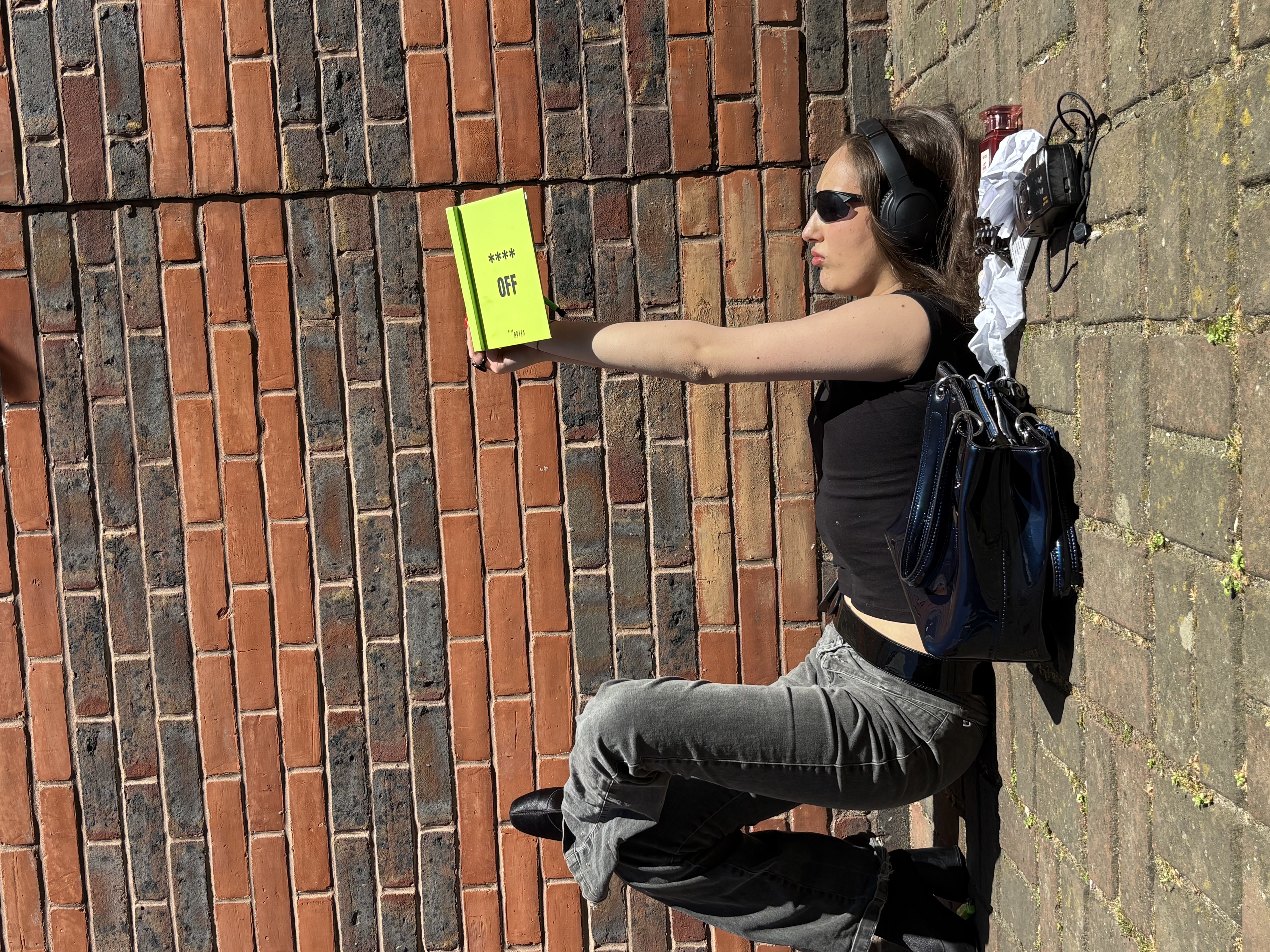 Green girl on a brick wall, headphones on, holding the **** OFF notebook