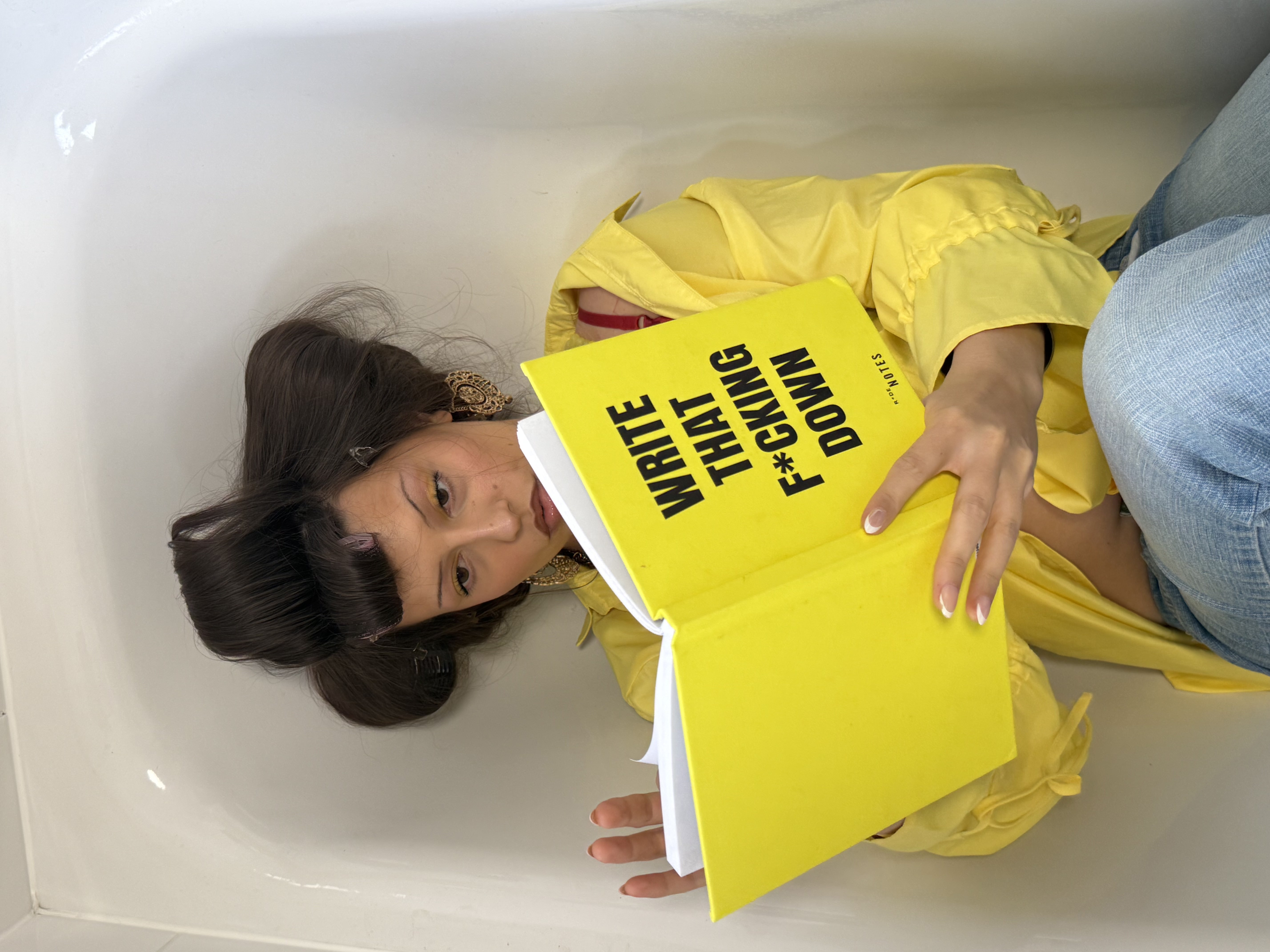 Yellow girl in a yellow shirt sitting in a bathtub holding the yellow WRITE THAT F*CKING DOWN notebook